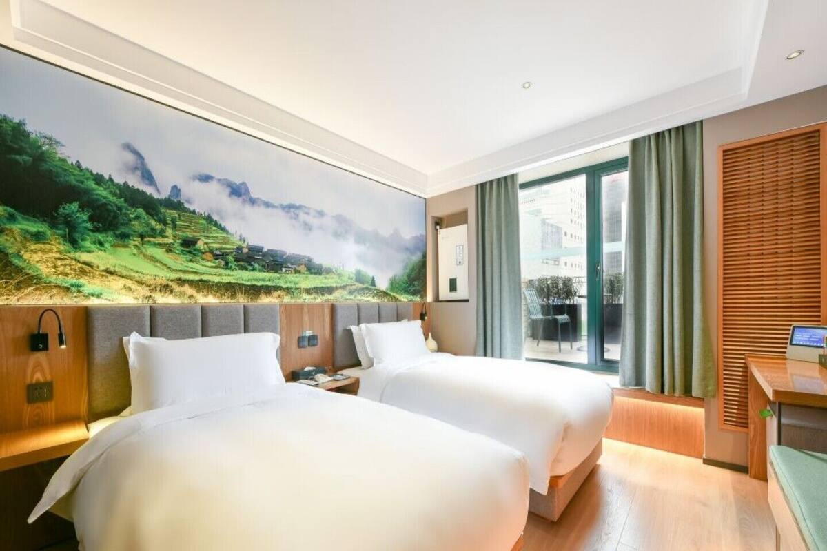 Habitación de hotel con dos camas, un ventanal con cortinas, un escritorio con computadora y un cuadro de un paisaje en la pared.
