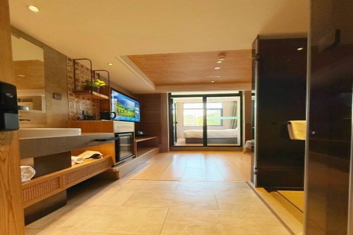 Una habitación de hotel moderna con un televisor grande, estanterías de madera, un cerramiento de vidrio corredero a un balcón y suelo de baldosas.