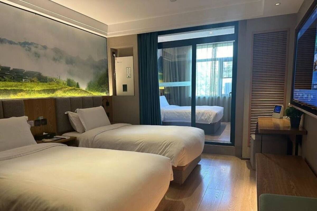 Habitación de hotel con dos camas, un escritorio, un televisor y un ventanal grande con cortinas.