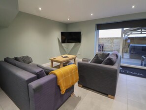 Ein modernes Wohnzimmer mit Sofa, Sesseln und einem kleinen Tisch. Ein Flachbildfernseher ist an der Wand befestigt.