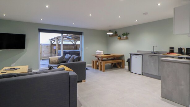 A modern living room with a grey sofa, a wooden dining table, and a kitchen area with a fridge and sink. - Oak Barn (Chester)