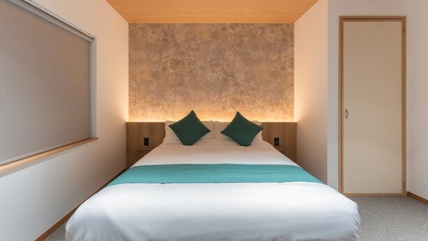 Een moderne hotelkamer met een groot bed, twee groene kussens, een blauwgroene plaid en een structuurwand achter het bed.