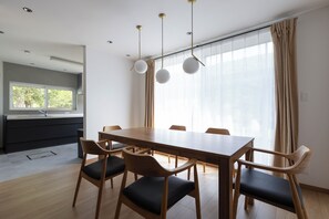 Ein moderner Essbereich mit einem Holztisch und -stühlen, im Hintergrund eine Küche und große Fenster mit Vorhängen.