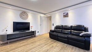 Een moderne woonkamer met een zwarte leren bank, een flatscreen televisie op een houten dressoir, een wandklok en een deur die naar een andere kamer leidt.