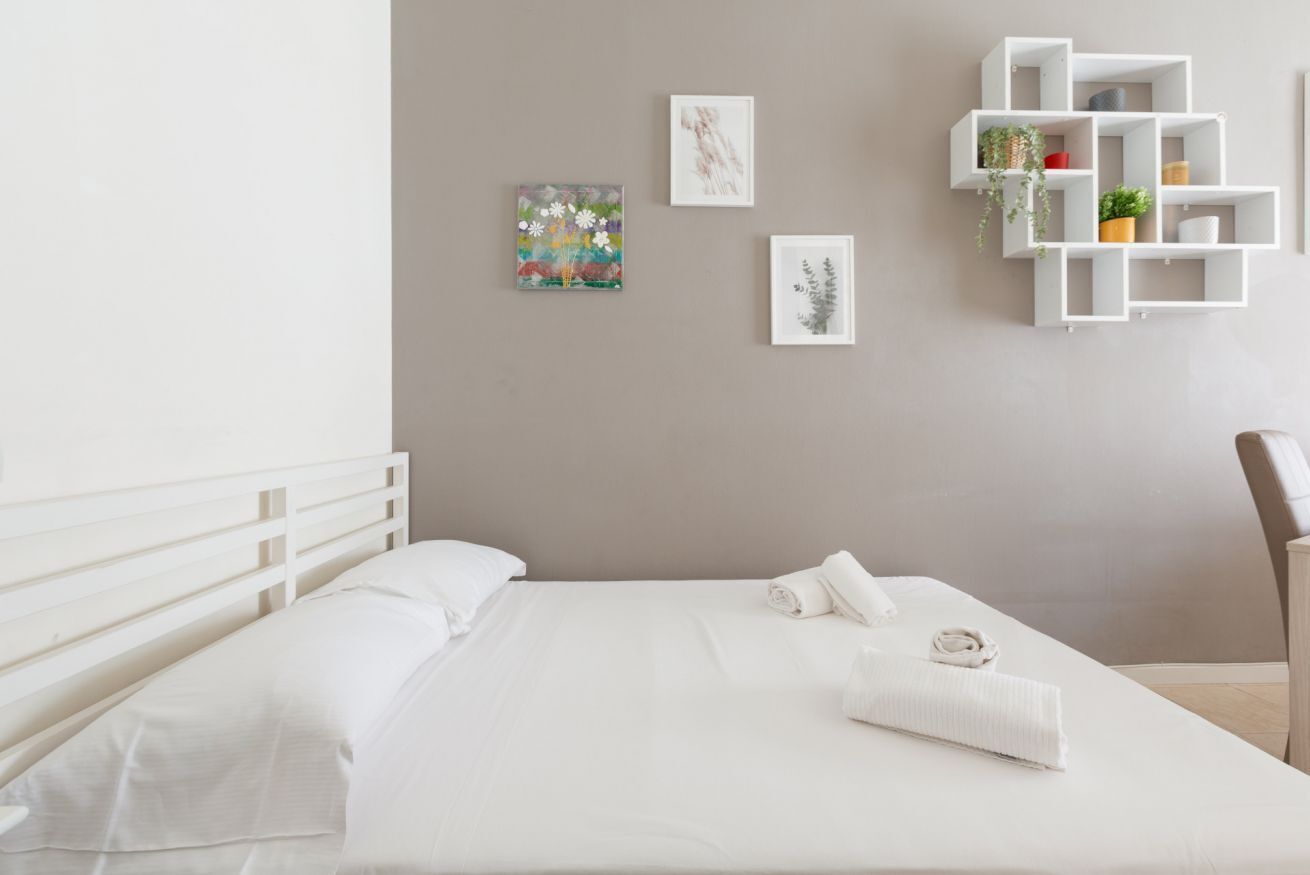 A neatly made bed with white linens, a white headboard, and a white bedside table with a lamp. There are three framed artworks on the wall and a white shelving unit with plants and decorative items.
