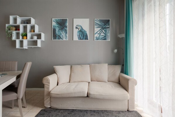 A living room with a sofa, a white shelving unit with plants, and three framed artworks on the wall.