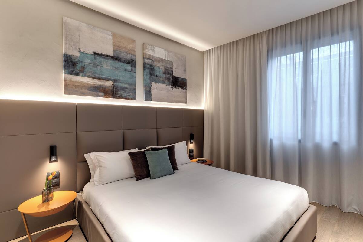 A modern hotel room with a large bed, two wall-mounted artworks, a bedside table with a lamp, and a large window with sheer curtains.