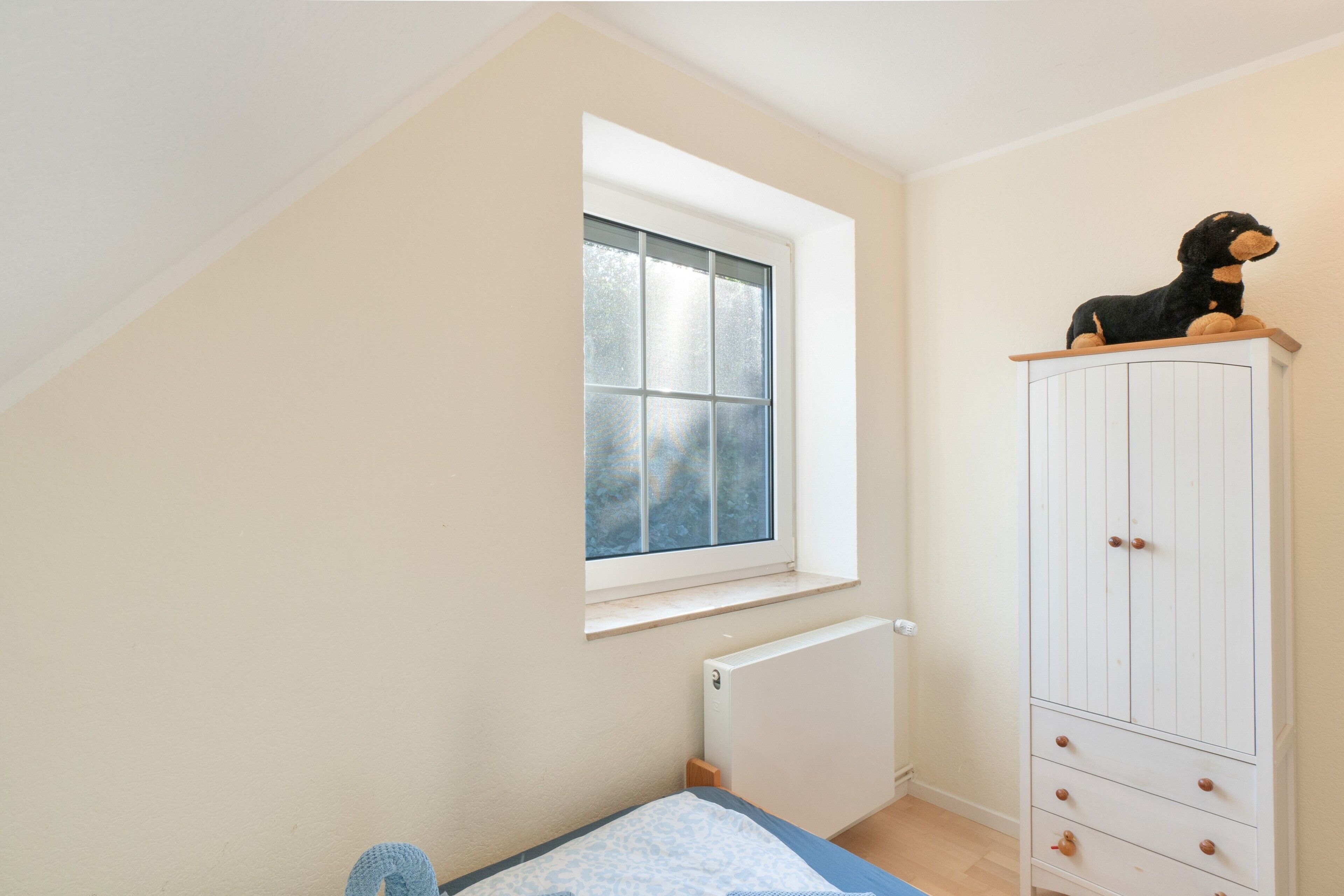 Ein Schlafzimmer mit schrägen Wänden, einem Fenster mit Milchglas, einem Heizkörper, einem Bett mit blumenmusterbezogener Decke in Blau und Weiß sowie einem weißen Kleiderschrank mit Holzgriffen.