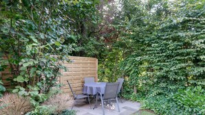 Eine Terrasse mit Tisch und Stühlen, umgeben von dichtem Grün und einem Holenzaun.