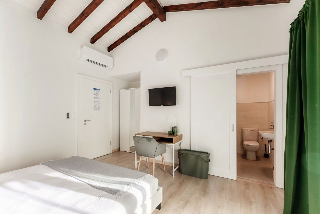 A bedroom with a bed, a desk, a chair, a television, and an open bathroom door.