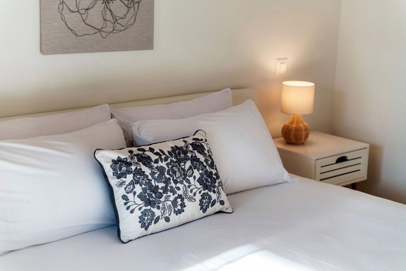 A neatly made bed with white bedding and a decorative pillow, a bedside lamp, and a small drawer unit.