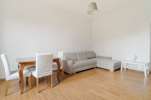 A modern living room with a wooden dining table, white chairs, a grey sofa, and a white side table.