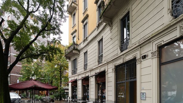 A building with multiple storefronts, outdoor seating, and a tree in the foreground. - GuestHost - Comfortable and bright 70m2 apartment, for 4 people, located on the 3rd floor of a building with elevator (NOT SUITABLE FOR DISABLED GUESTS). The property is located in the Isola neighborhood, very young and full of services, well served by p (Milan)