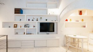 Una sala de estar moderna con televisor empotrado, estanterías blancas y un comedor con mesa y sillas blancas.