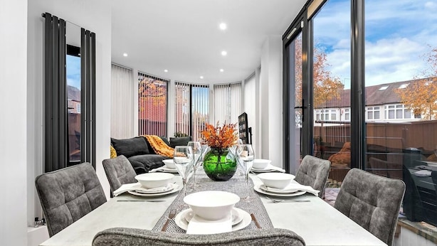 A modern dining area with a rectangular table set for six, surrounded by grey upholstered chairs. A green vase with orange flowers is placed at the center of the table. - Modern 4BR London Home with Games Room Large Garden Free Parking and Fast WiFi (Wembley)