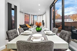 A modern dining area with a rectangular table set for six, surrounded by grey upholstered chairs. A green vase with orange flowers is placed at the center of the table. - Modern 4BR London Home with Games Room Large Garden Free Parking and Fast WiFi (Wembley)