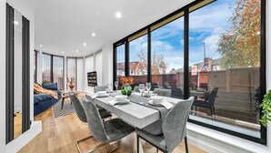 A modern dining area with a large window, a sofa, and a dining table set for four. - Modern 4BR London Home with Games Room Large Garden Free Parking and Fast WiFi (Wembley)
