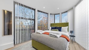 A modern bedroom with a large bed, a green headboard, and a view through a window with a view of trees and buildings. - Modern 4BR London Home with Games Room Large Garden Free Parking and Fast WiFi (Wembley)