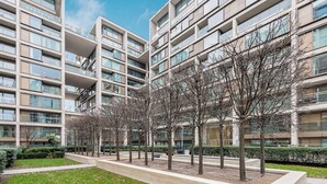 Un immeuble résidentiel moderne à plusieurs étages, doté de balcons, d’une cour aménagée avec des arbres et d’une pelouse bien entretenue.