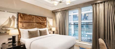 A modern hotel room with a large bed, a wooden headboard, a nightstand, a lamp, a blue rug, and a view of the city through large windows.