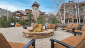 A stone fire pit surrounded by outdoor seating, a clock tower, and lodge-style buildings in a mountainous area. - Prime Location, Steps from the Ski Lift - 2 Bedroom Condo in Arrowhead Village 2 Condo (Edwards)