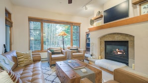 A living room with a fireplace, leather sofa, and a large window with a view of trees. - Prime Location, Steps from the Ski Lift - 2 Bedroom Condo in Arrowhead Village 2 Condo (Edwards)
