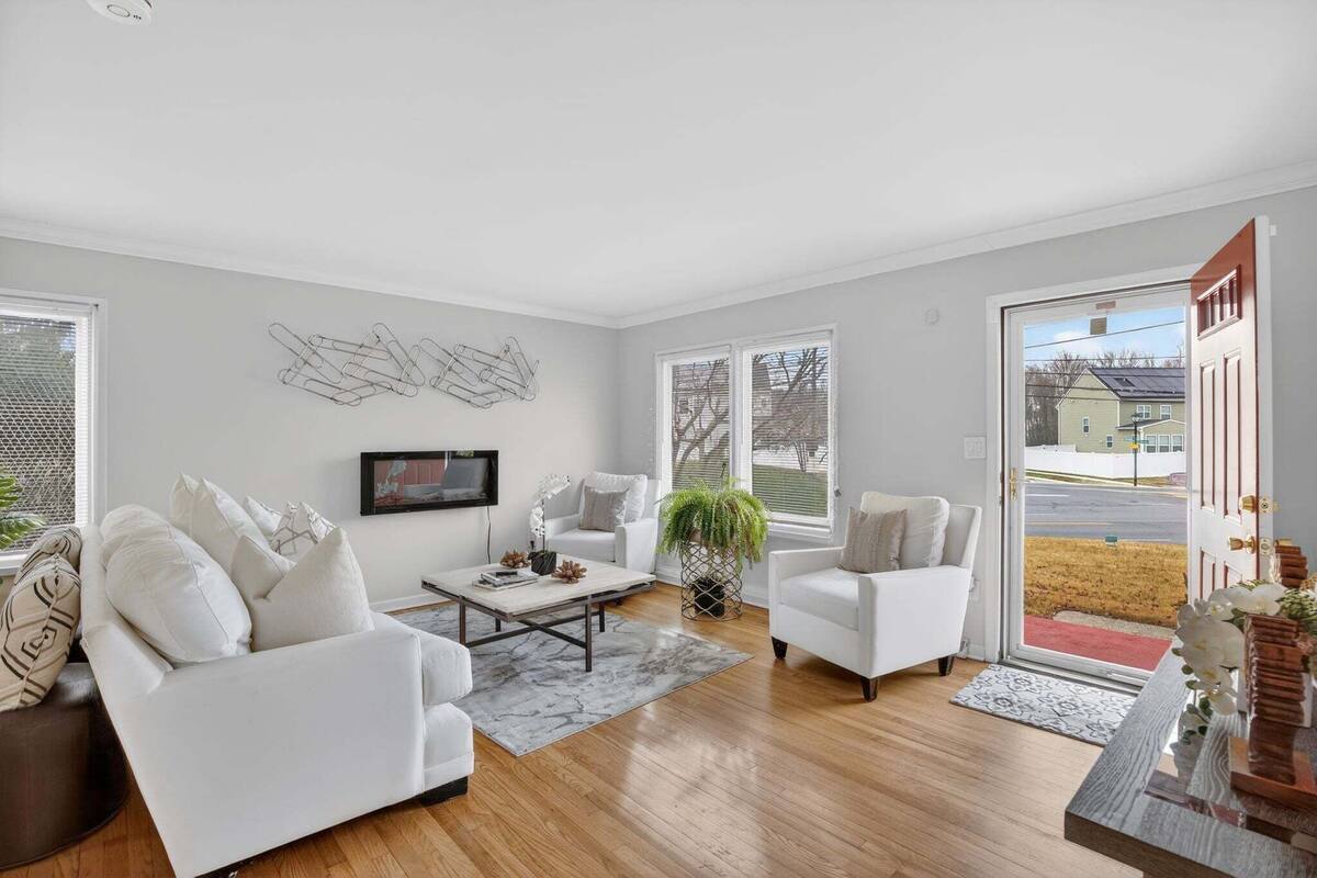 Un salon spacieux avec un canapé blanc, une table basse et un téléviseur fixé au mur. Une grande fenêtre offre une vue sur l’extérieur.