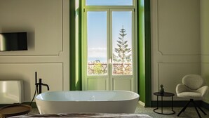 A modern bathroom with a freestanding bathtub, a television mounted on the wall, and a view of a tree through a window. - Bright and Elegant Sea-View Room with a Timeless Charm (Acireale)