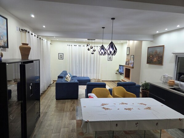 A modern living room with a sofa set, a dining table, and a kitchen area with a refrigerator.