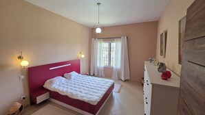 A bedroom with a red headboard, a bed with white bedding, a bedside table, a dresser, and a window with curtains.