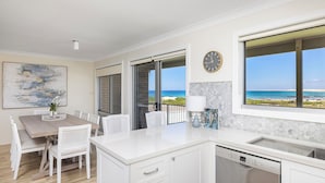 A modern kitchen with a view of the beach, a dining area with white chairs, and a clock on the wall. - Absolute Birubi Beachfront - Luxury Pet Friendly (Anna Bay)