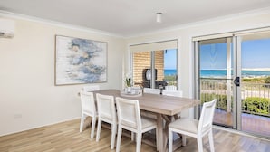A dining area with a wooden table and white chairs, a painting on the wall, and a view of the sea through a sliding glass door. - Absolute Birubi Beachfront - Luxury Pet Friendly (Anna Bay)