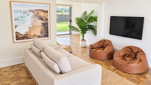 A living room with a sofa, two bean bags, a potted plant, and a TV mounted on a white brick wall. - Absolute Birubi Beachfront - Luxury Pet Friendly (Anna Bay)