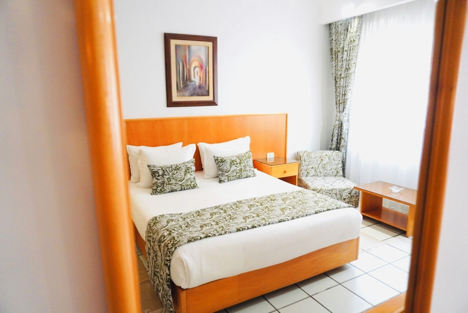A hotel room with a wooden bed, a nightstand, a chair, and a painting on the wall.