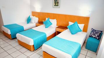 A hotel room with four single beds, each with a wooden headboard, white bedding, and blue throw pillows. There is a small bedside table with a drawer on each bed.