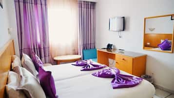 Quarto de hotel com cama, escrivaninha, cadeira, televisão e espelho.