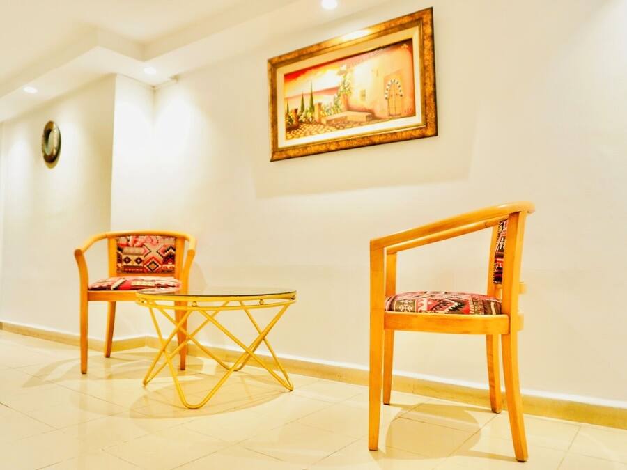 A hotel lobby with two wooden chairs with patterned cushions, a small round table, and a framed painting on the wall.