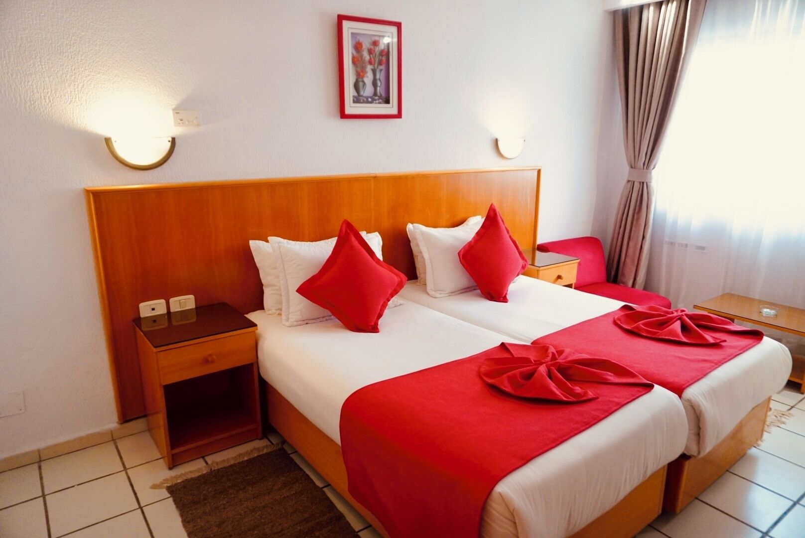 Une chambre d’hôtel avec deux lits, une literie rouge et blanche, une tête de lit en bois, des tables de chevet et des lampes fixées au mur.