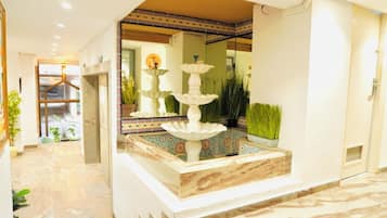 Zona de recepción con piso de mármol, una fuente de tres niveles y un espejo que refleja el espacio.