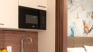 A modern kitchen with a built-in microwave, a wooden backsplash, and a sink with a modern faucet. There is a fruit bowl on the counter and a bed with white bedding in the adjacent room. - OLIVER pulse I Self check-in Apartments (Vienna)