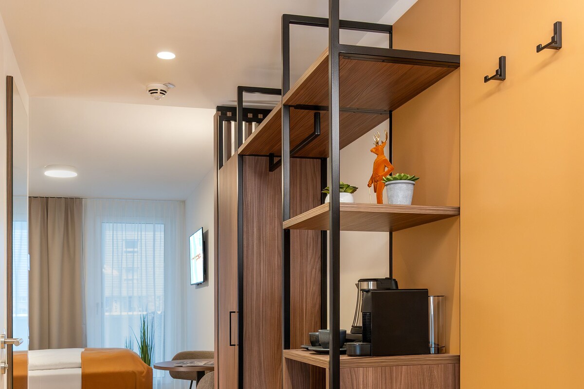 A modern hotel room with a wooden floor, a bed with orange bedding, a desk with a computer, and a coffee machine.