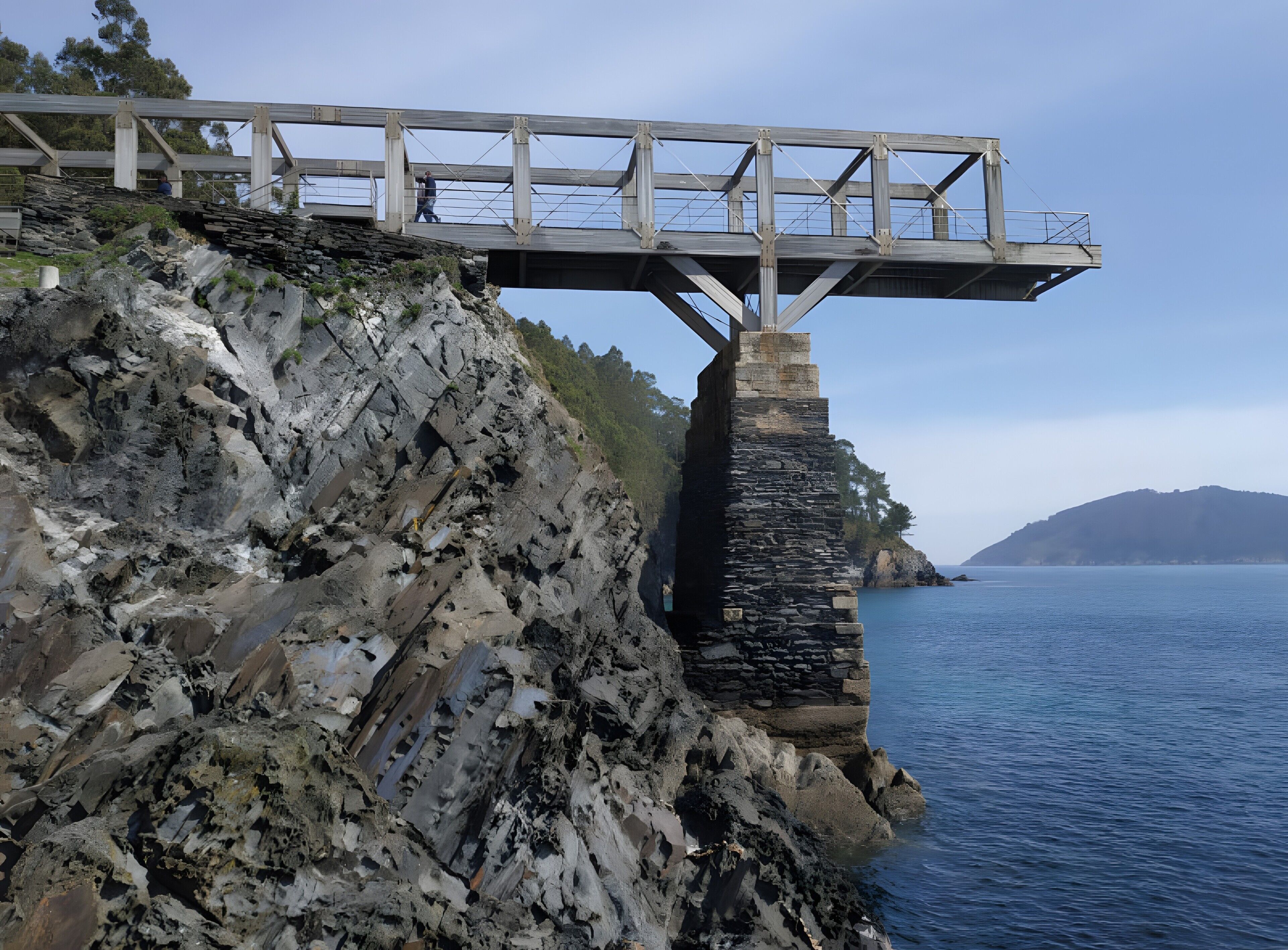 Un pont enjambant une falaise rocheuse, offrant une vue sur la mer et les collines lointaines.