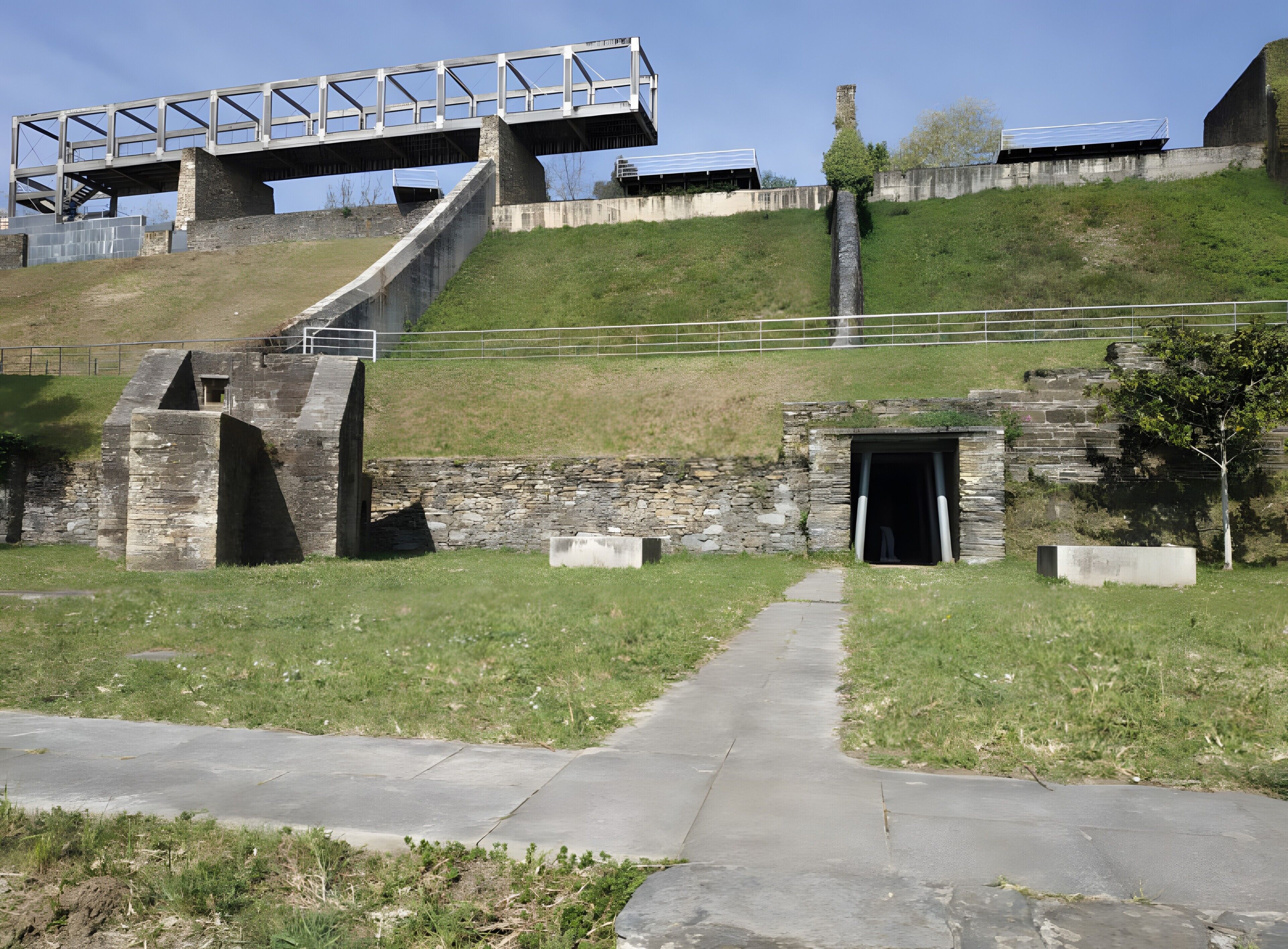 Un sendero de hormigón que conduce a una construcción de piedra con una escalera de metal y un puente encima.