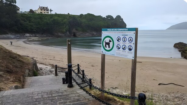 Uma praia com placa de uso compartilhado, cerca de arame e uma escadaria de pedra que desce até a praia.