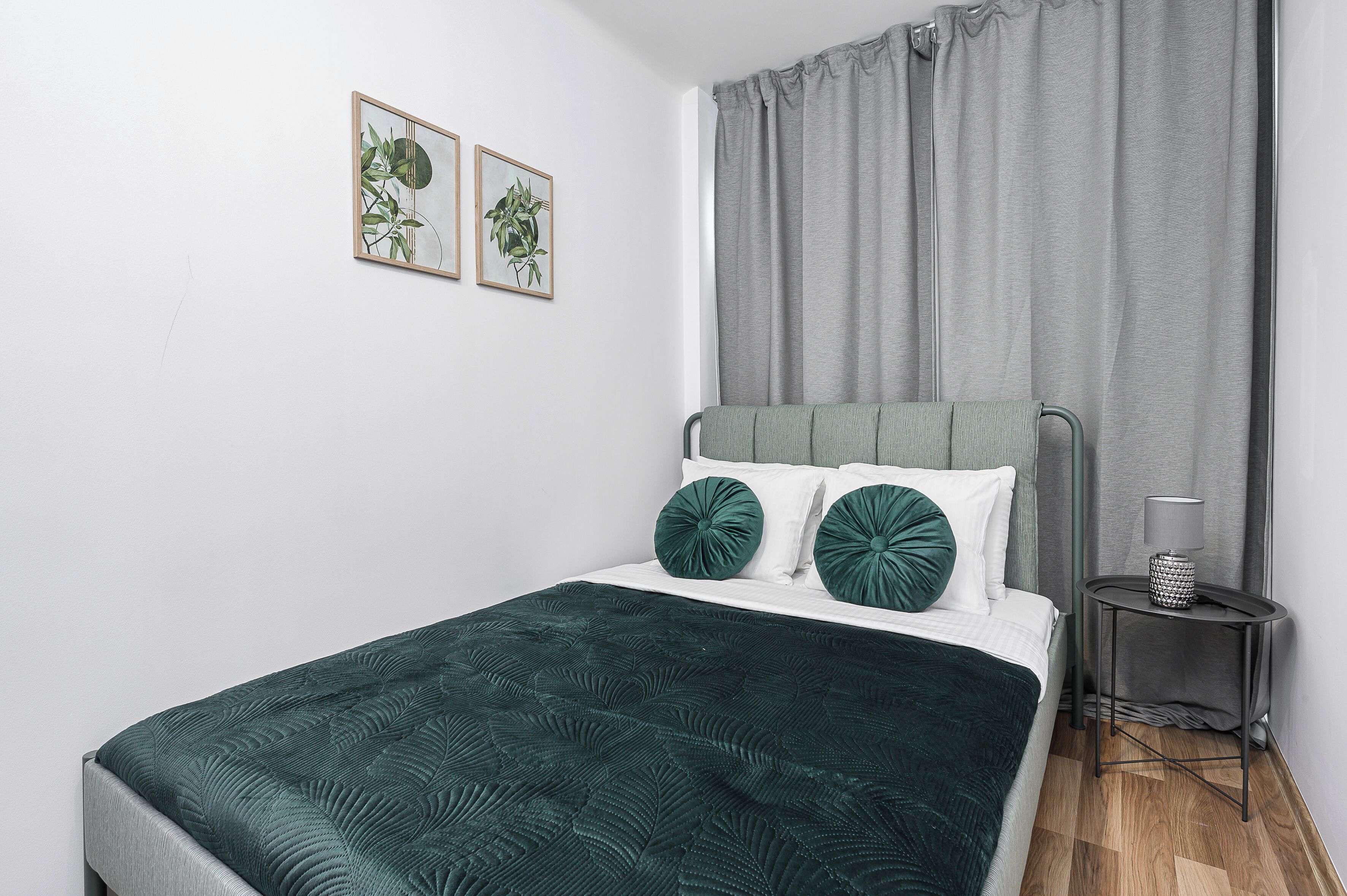 Een net opgemaakt bed met een groene sprei en bijpassende kussens, een grijs hoofdbord en een klein nachtkastje met een lamp.