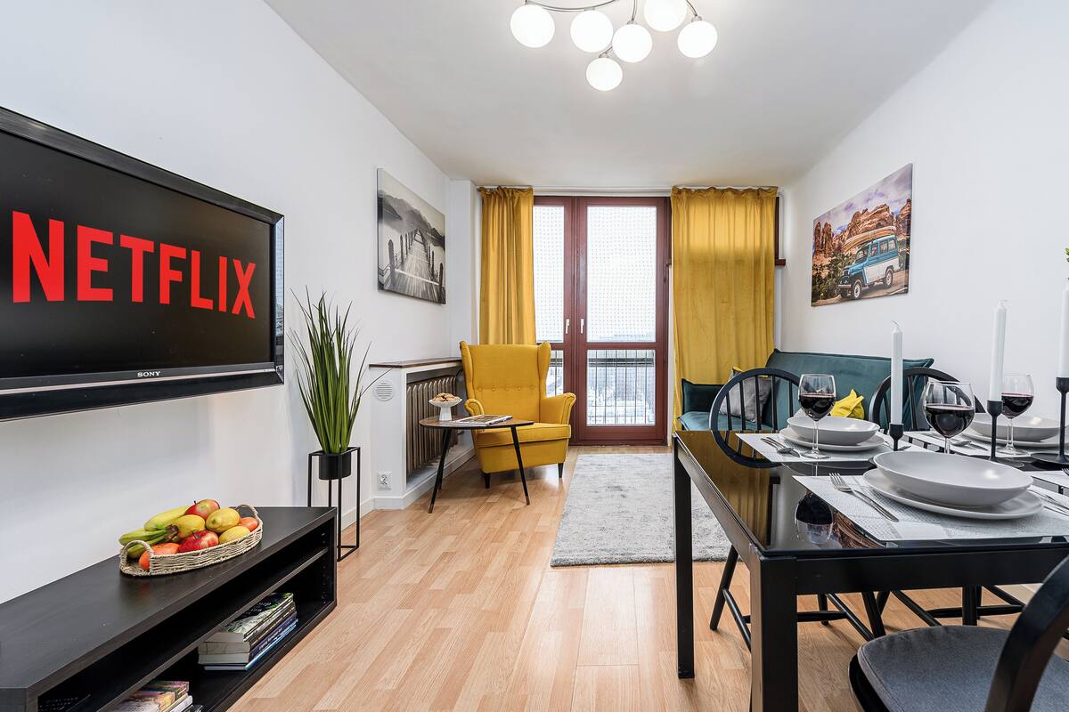 Una sala de estar moderna con un televisor de pantalla plana mostrando el logotipo de Netflix, un comedor con una mesa lista para dos personas y un sillón amarillo.