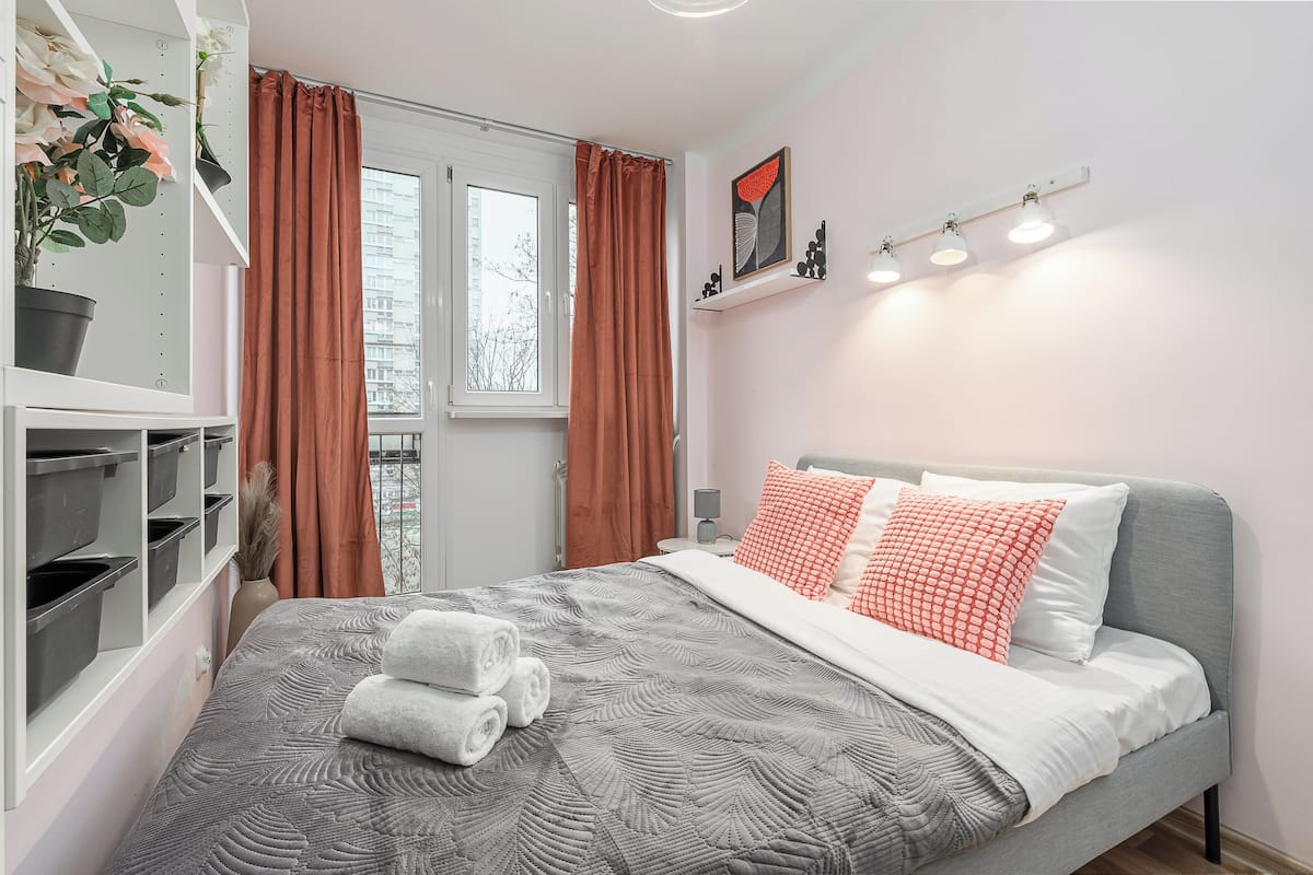 Una cama bien hecha con una colcha gris estampada, almohadas blancas y una almohada roja y blanca. Hay dos plantas en macetas sobre una estantería, una ventana con cortinas y un aplique de pared.