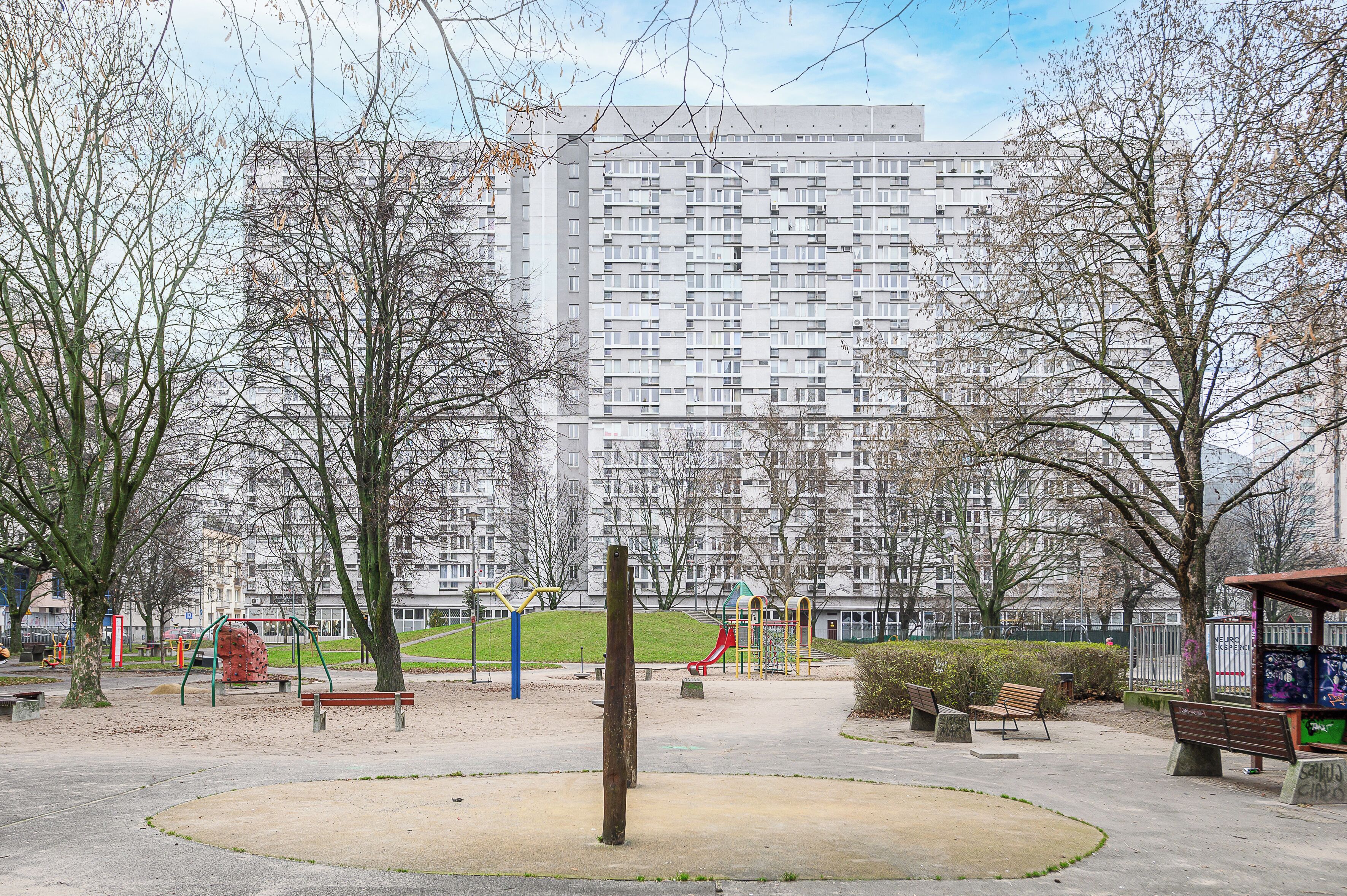 Um parque com equipamentos de recreação, bancos e um prédio grande ao fundo.