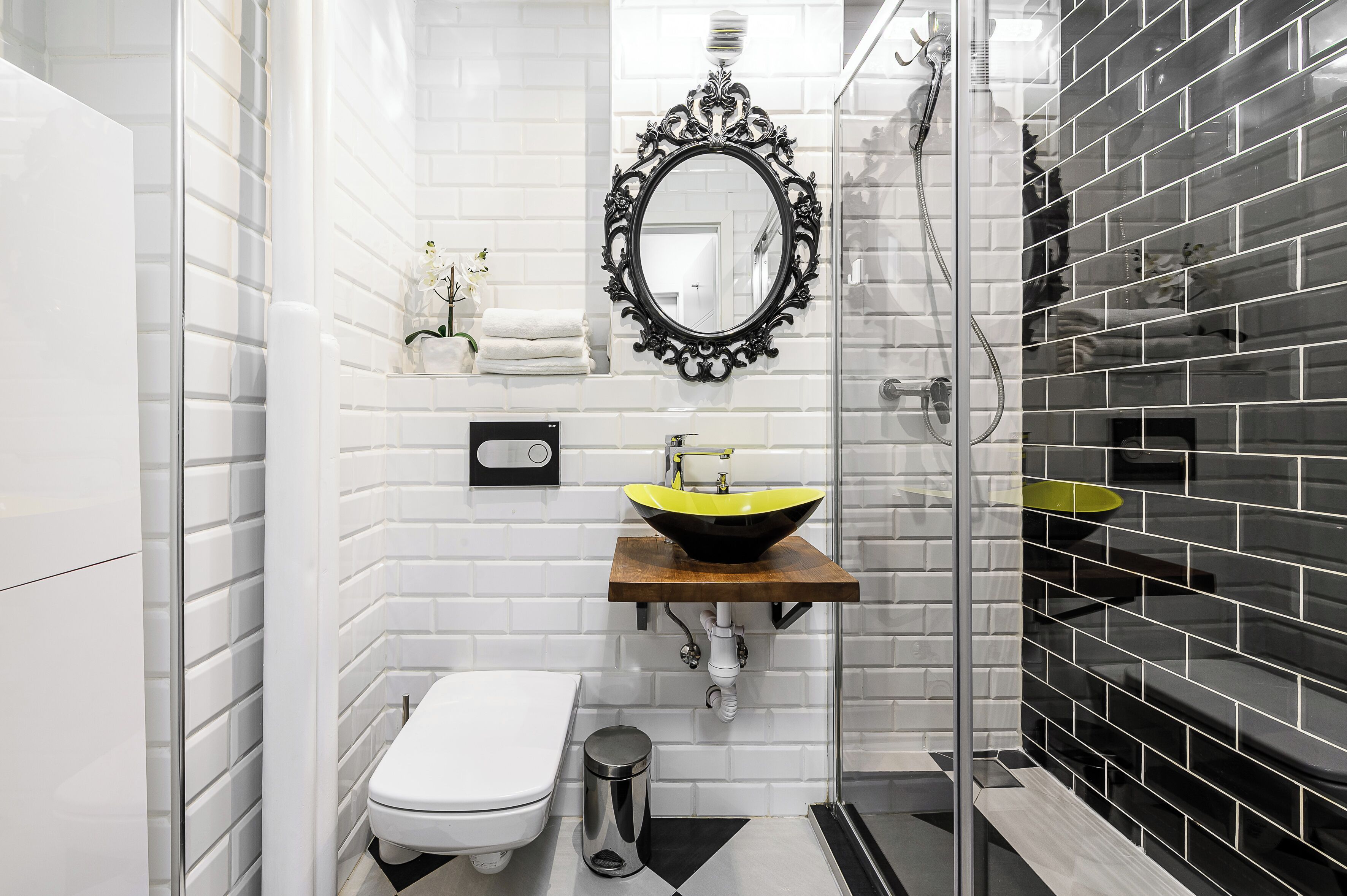 Ein modernes Badezimmer mit weiß gefliesten Wänden, einem spiegel mit schwarzem Rahmen, einem gelben Waschbecken, einer weißen Toilette und einer Duschkabine aus Glas.