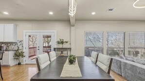 Uma sala de jantar com mesa de madeira escura, cadeiras estofadas brancas e uma planta central. Há janelas amplas com persianas, uma porta branca e vista para uma varanda.